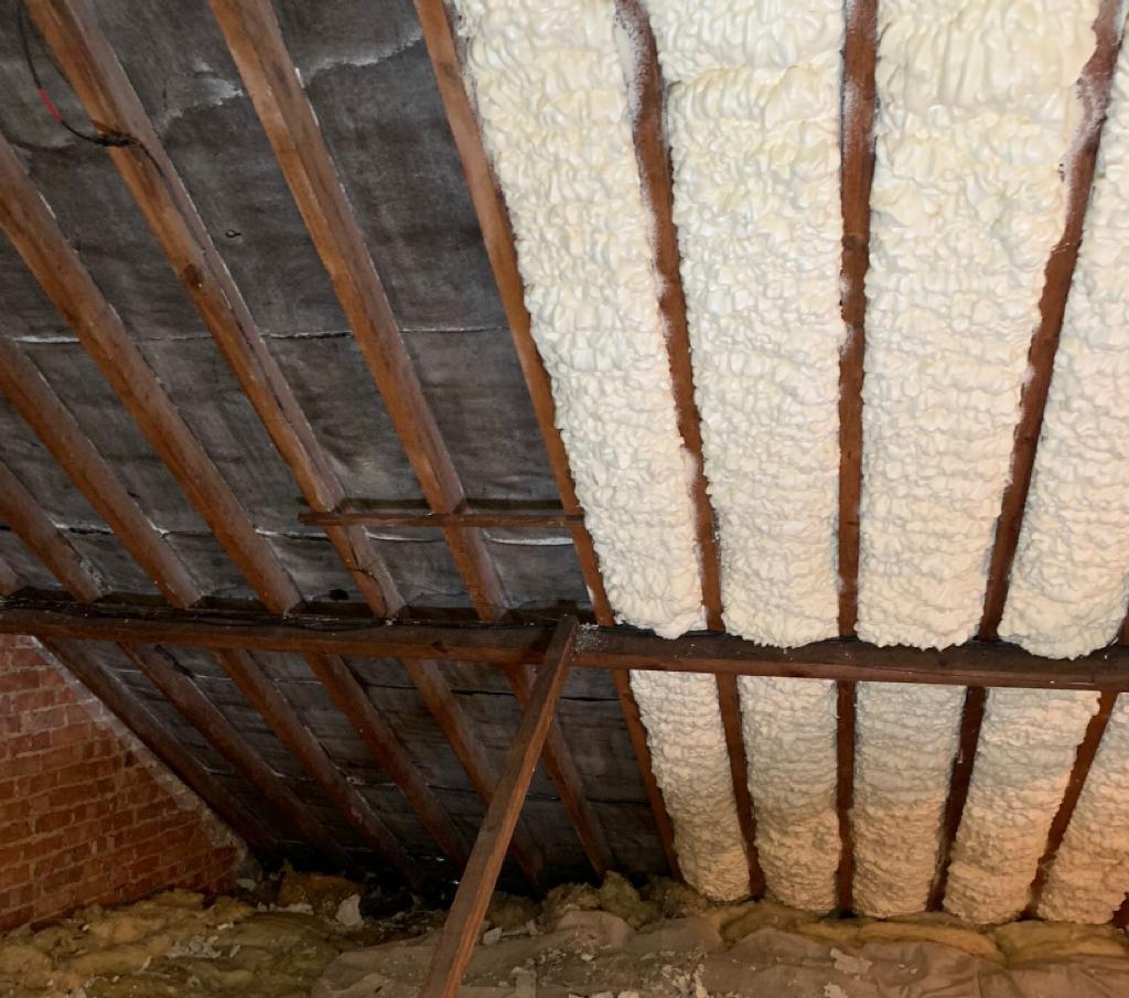 The image shows the interior of an attic or a similar space under a roof. You can see wooden rafters or trusses that support the roof, and between them, there is sprayed foam insulation which has expanded and solidified. This is a common method used to insulate buildings to maintain temperature and reduce energy consumption. The darker wood and the bricks suggest that it might be an older building. Some of the rafters appear to have not been sprayed yet, as they still show the bare wood. There is also a brick wall visible, indicating that this could be the top part of an exterior wall, or it could be a chimney. The floor of the attic is not fully visible, but there looks to be some debris and possibly older, less effective forms of insulation material, like mineral wool or cellulose, that might be replaced or supplemented by the spray foam.