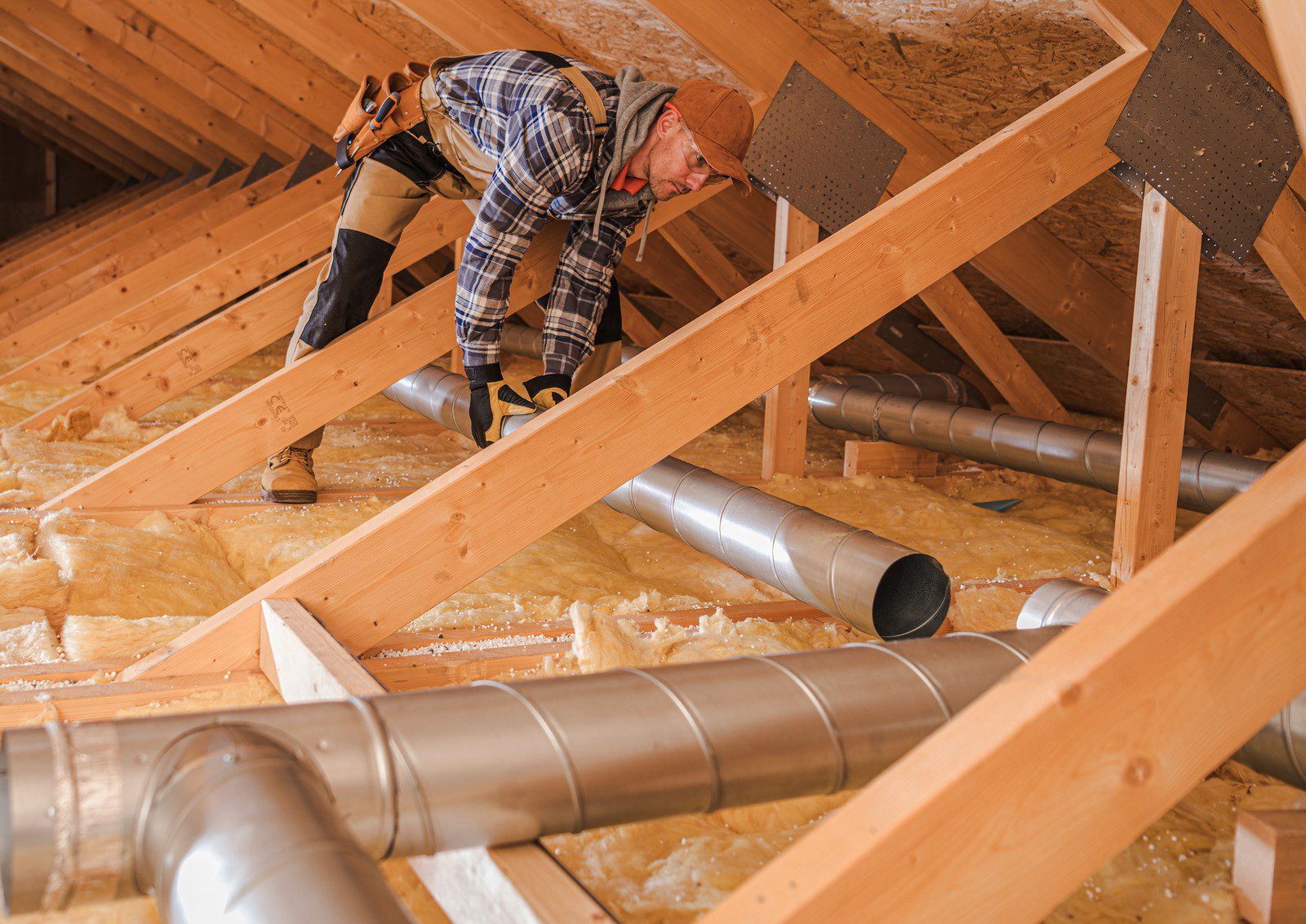 The image depicts a person working in an attic space. The individual is wearing a work outfit with tools attached to their belt, protective gloves, and a cap. They appear to be engaged in a task related to the construction or maintenance of the attic, possibly installing or inspecting ventilation ducts.

The attic space features wooden rafters and beams, and there are insulation materials spread across the floor. Several metal ducts run through the area, indicating that some form of ventilation or air conditioning system is being implemented or serviced.

The roofing material is visible from the inside, and vented soffit material is installed between the rafters to promote airflow. The worker is carefully stepping on the wooden beams to avoid damaging the insulation or falling through the ceiling below. This setting suggests that the work being done may be part of a home improvement, renovation, or energy efficiency project.