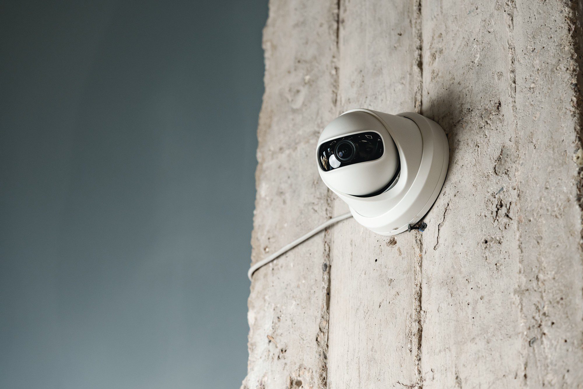 The image shows a modern, white dome security camera mounted on a concrete wall. The camera appears to have infrared capabilities for night vision, as suggested by the visible LEDs surrounding the lens. There is a cable connected to the camera that runs along the wall, likely providing power and possibly network connectivity. The background is a textured concrete surface, and the colour scheme of the photo is neutral with the camera as the main focus.