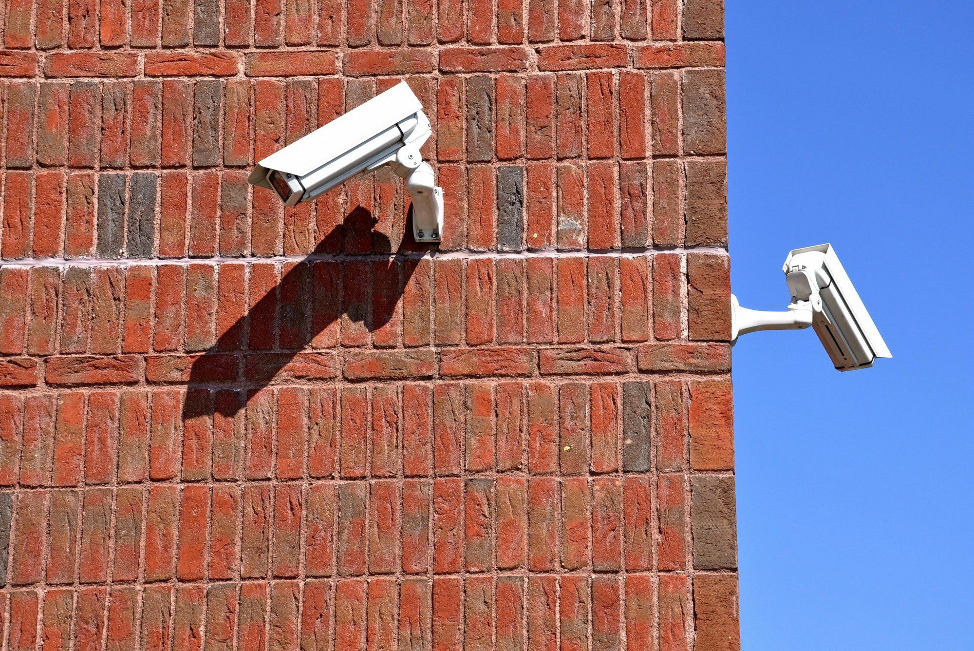 The image shows two security cameras mounted on a red brick wall, with a clear blue sky in the background. The sunlight casts shadows of the cameras on the wall, emphasizing their presence. Each camera is positioned at a corner of the wall, allowing for surveillance from different angles. The scene suggests a focus on security and monitoring.