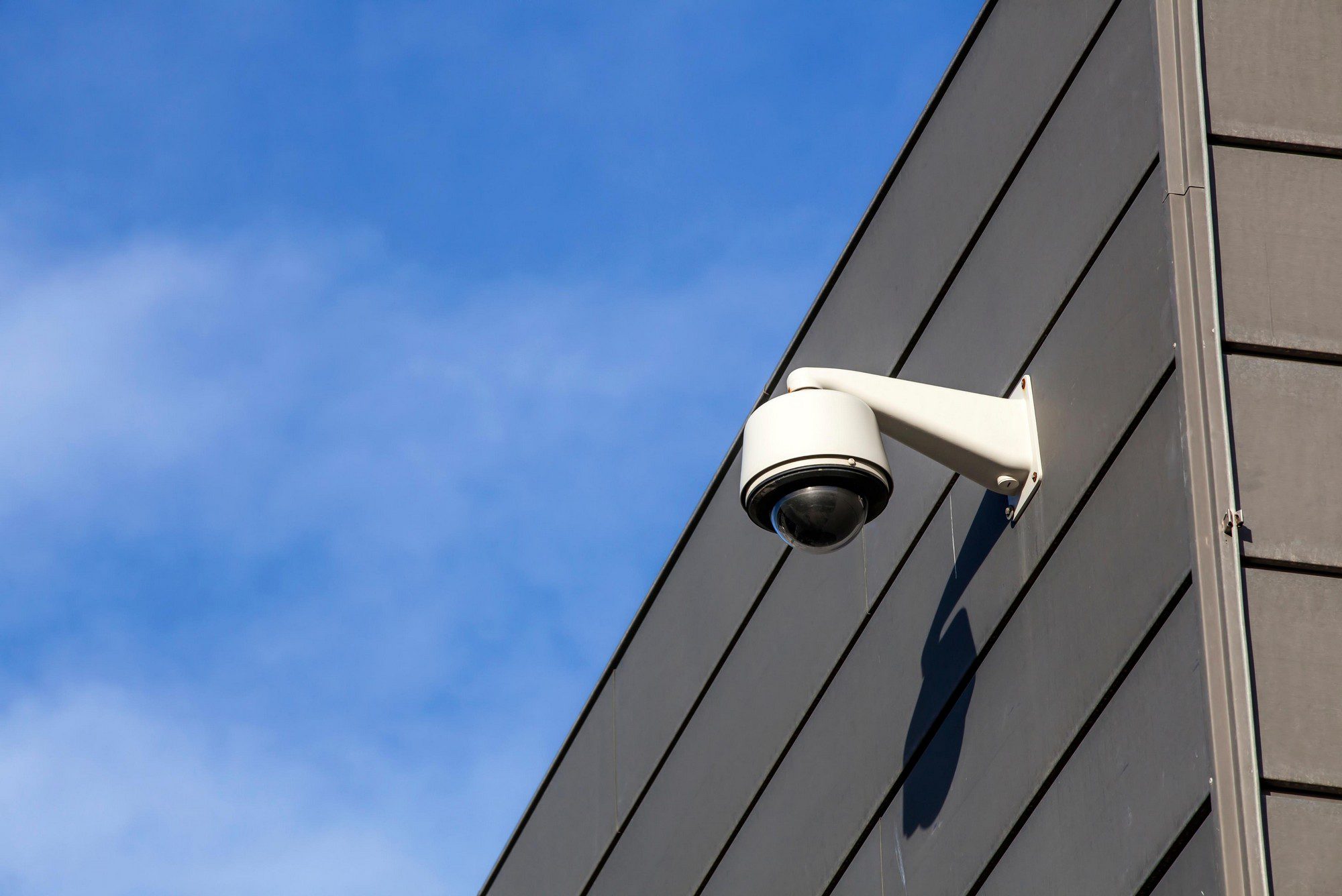 The image shows a security camera mounted on the exterior wall of a building. The camera has a protective dome cover and is positioned to overlook the area around the building. The backdrop is a clear sky. The camera is likely used for surveillance to enhance security by monitoring and recording activities in its field of view.