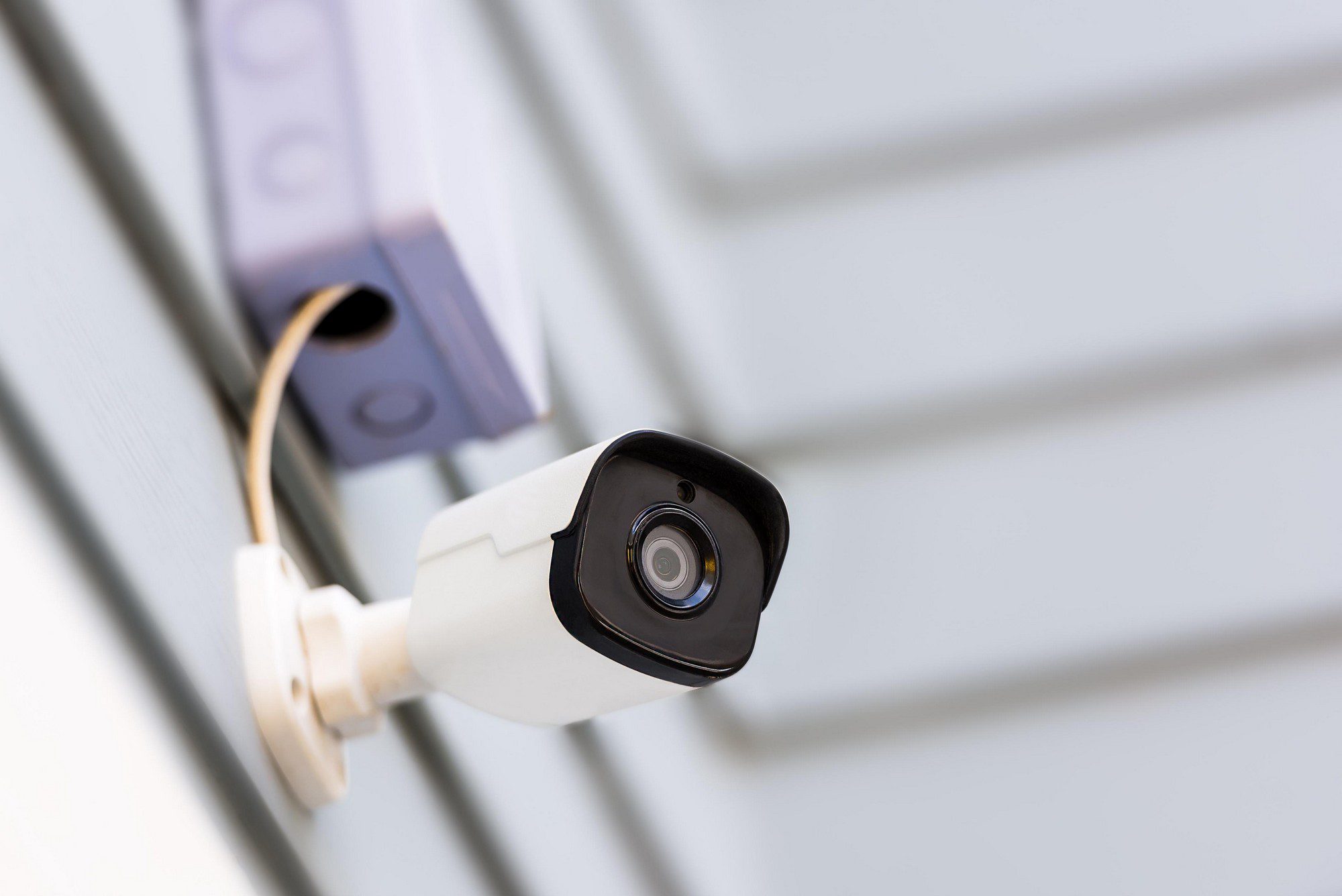 The image shows a modern security camera mounted on a wall. The camera has a black face with what appears to be a lens in the centre, indicative of a device used for surveillance or monitoring purposes. The wiring of the camera is partially visible, as it runs from the back of the camera into the wall, suggesting a hardwired power connection. The background is a light-coloured, vertically grooved surface which could be siding on the exterior of a building. The focus of the image is on the camera, highlighting its design and position.