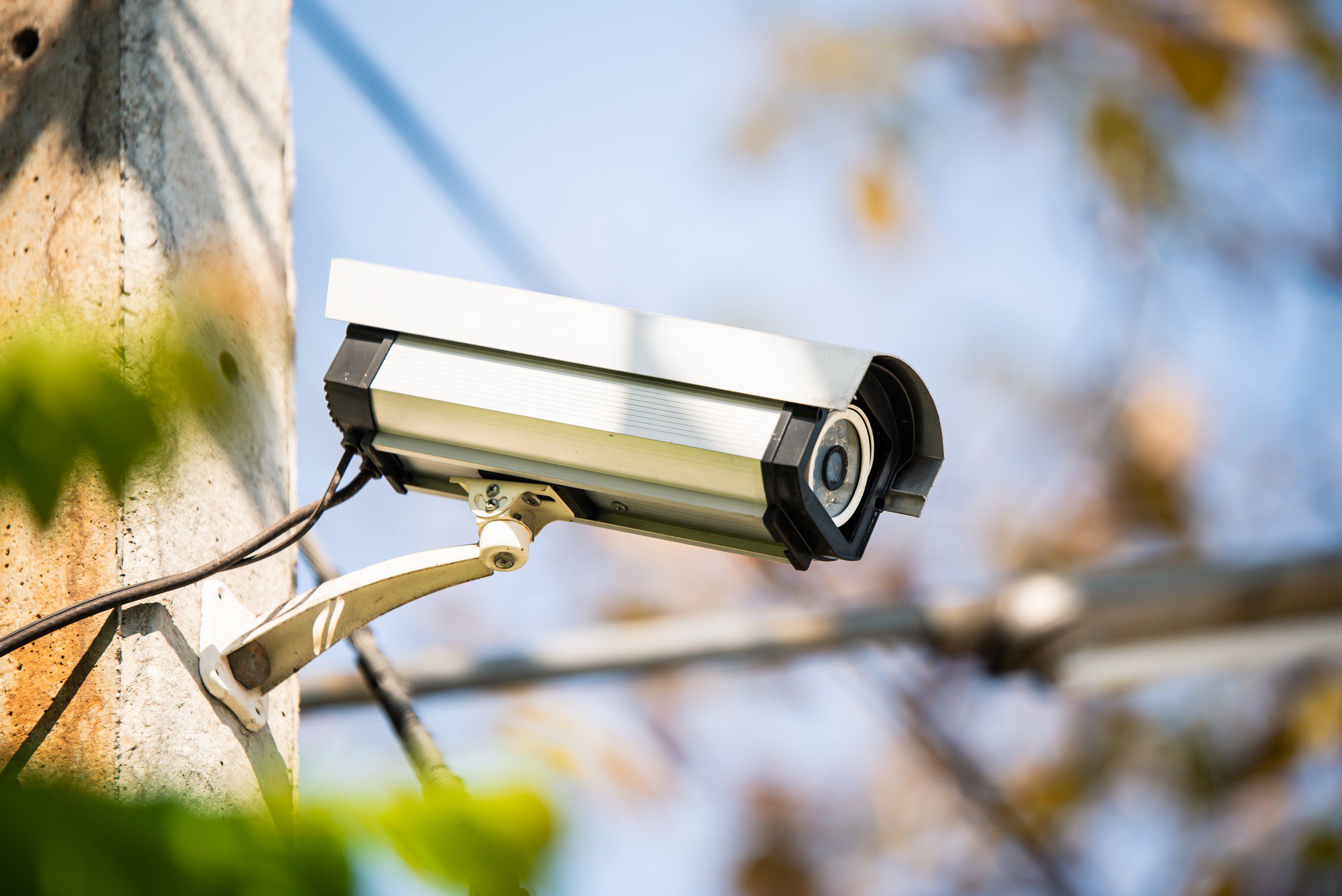 This image shows an outdoor security camera mounted on a pole. The camera is encased in a protective white housing and is positioned to surveil the surrounding area. In the background, there are blurred leaves and branches, suggesting the camera is installed in a location with some vegetation, perhaps a park or an area around a building with greenery. The wires are visible, indicating the camera is connected to a power source and possibly to a monitoring system. The image captures a clear day with good visibility for the camera's operation.