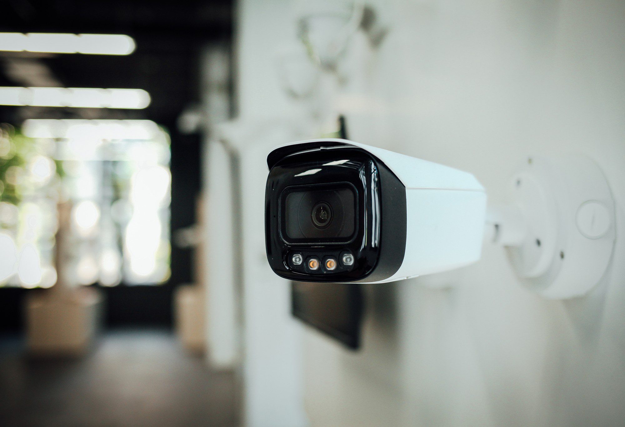 The image shows a close-up view of a modern CCTV security camera mounted on a wall. The camera is positioned off-centre to the right side of the frame and is white with a black camera lens area. It appears to be an outdoor-type camera given its sturdy design and protective casing. Below the main lens, there are additional smaller sensors or lights, which could be infrared LEDs for night vision. The background is a bit blurred due to the shallow depth of field, emphasising the camera in the foreground. The interior setting has a corridor-like appearance with what looks like a diffuse daylight coming through, illuminating the scene.