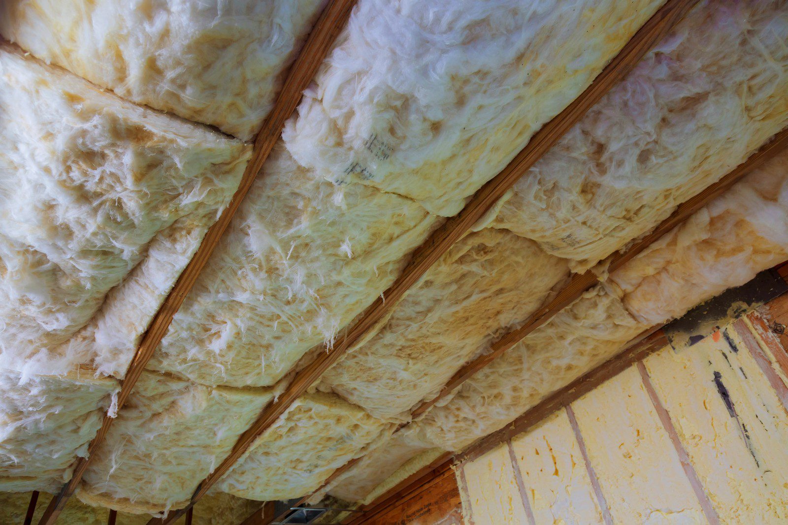 This image shows the underside of a roof with fiberglass insulation installed between the wooden rafters. The insulation appears to be unfaced fiberglass batts, which are a common material used to insulate buildings to improve energy efficiency by reducing heat transfer. At the bottom of the image, there's a partial view of a wall with what seems to be foam board insulation, identified by its yellow colour and rigid appearance. This setup is typical in attics or unfinished areas of a structure where the insulation is exposed rather than being covered with drywall or other finishing materials.