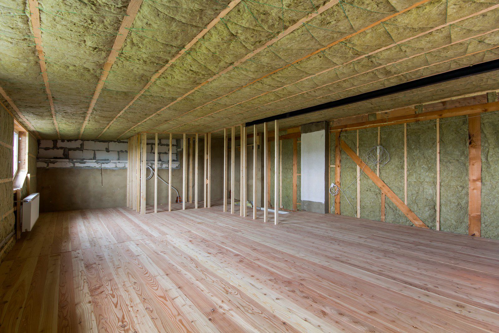 This image shows the interior of a room under construction or renovation. The room has exposed wall studs and visible insulation material in the walls and ceiling. The insulation appears to be fiberglass, recognizable by its fluffy, wool-like texture and yellowish colour. Wooden floorboards are laid down, covering the floor, and it looks like new hardwood flooring that has not yet been finished or sealed. There is a partial view of a cinder block wall on the back left side, indicating that part of the building may be below ground level or designed with a combination of materials for foundational support. Electrical wiring and conduit can be seen running through the wall studs, suggesting that electrical rough-in work has been started. A radiator or heating unit is present on the left side against the wall. The overall state of the room shows that it is in the process of being built or remodeled, with further work required for insulation coverage, electrical and plumbing installations, wallboard or drywall application, and finishing touches.