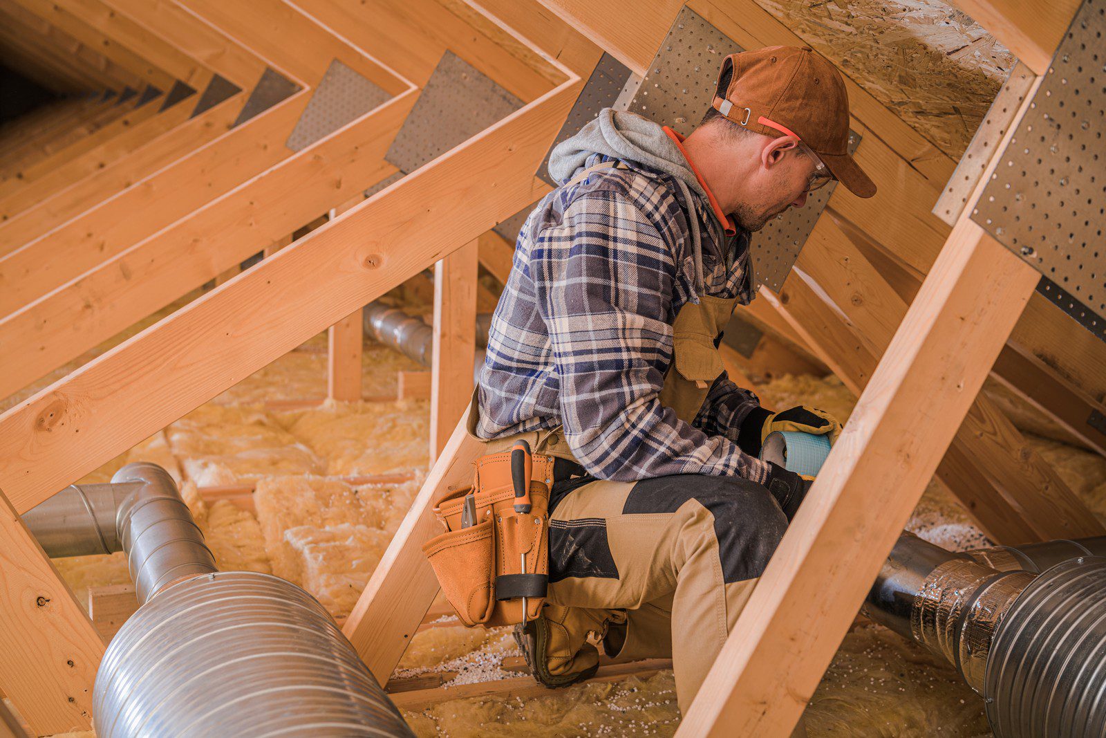 This image shows a person working in an attic space. The individual is wearing a plaid shirt, a hat, safety glasses, and a tool belt. They appear to be sitting on wooden beams surrounded by insulation material, which is often used to improve energy efficiency by sealing in heat. There are also ventilation ducts made of flexible material running through the attic space. The worker is likely engaged in some form of construction, maintenance, or installation work, possibly related to the HVAC (heating, ventilation, and air conditioning) system, given the presence of the ductwork.