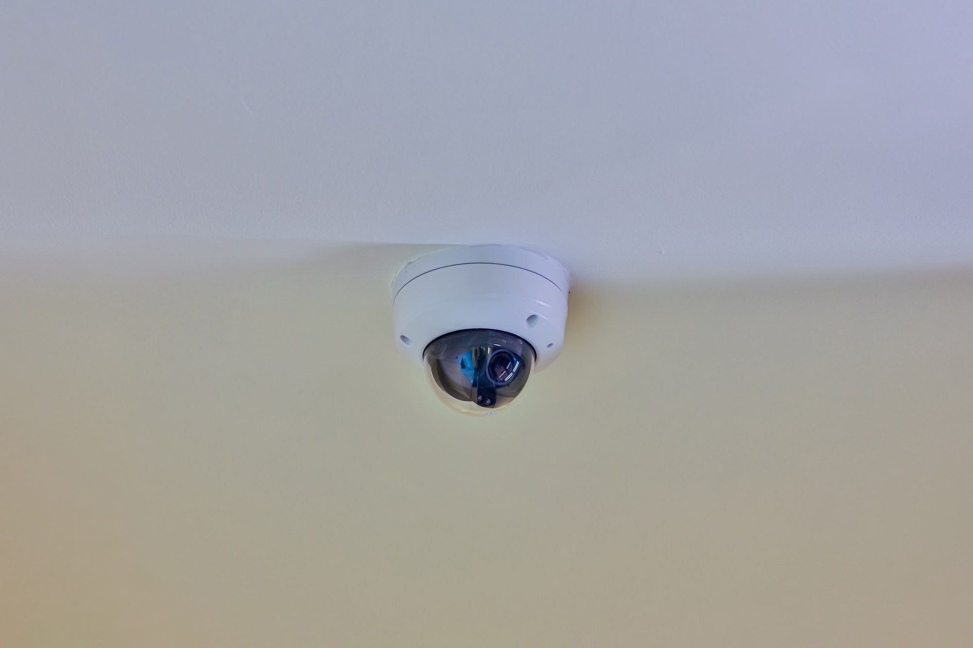 The image shows a dome security camera mounted on a ceiling. The camera is white and is facing downwards, which is a typical installation to monitor activities below. The wall and ceiling appear to be painted in light tones, providing a neutral background that emphasizes the presence of the camera.