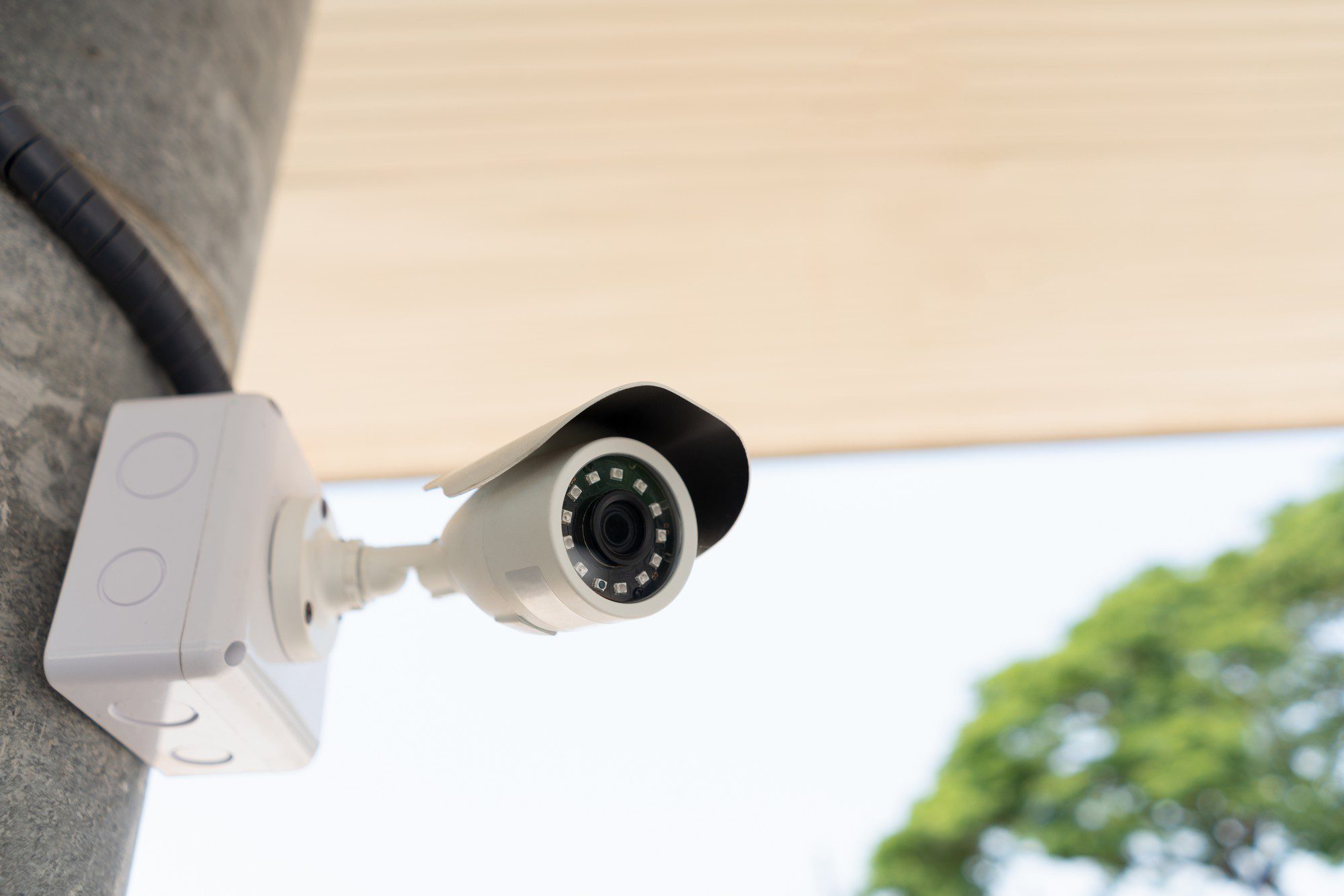 The image shows an outdoor security camera mounted on the exterior of a building. The camera is facing downwards, suggesting that it is monitoring the area below. The device appears to be a modern CCTV camera, equipped with infrared LEDs around the lens, which are typically used for night vision capabilities. A cable attached to the camera indicates it is connected to a power source or a recording system. In the background, there is a blurred view of a light-coloured ceiling or overhang, a piece of conduit, and some foliage from nearby trees, indicating an outdoor setting.