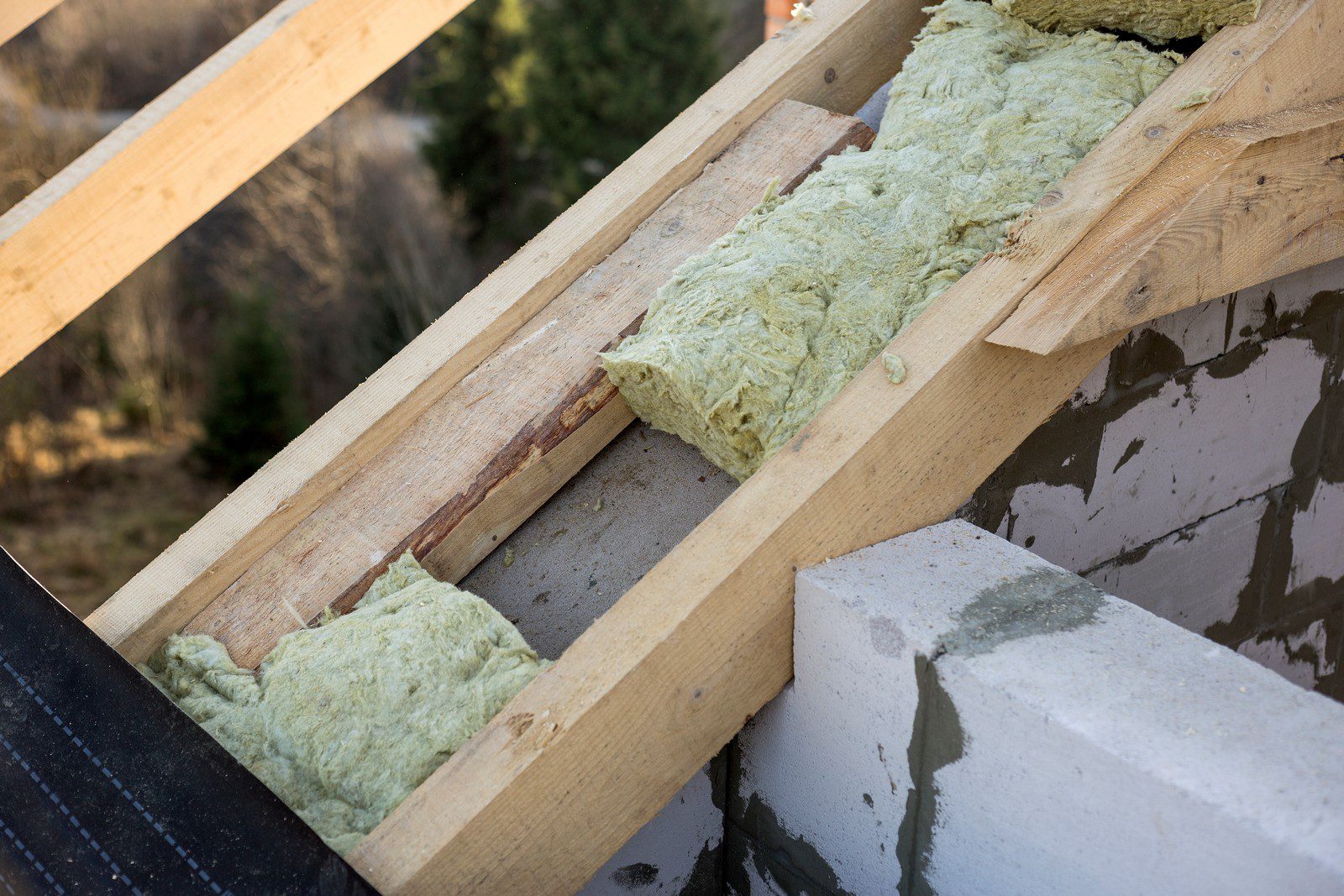 This image shows a construction or renovation scene where insulation material is being installed between wooden rafters. The greenish material appears to be fiberglass insulation, which is commonly used in buildings for thermal regulation and soundproofing. The wooden beams suggest that this could be part of a roof or ceiling structure. Additionally, there are concrete blocks visible, which might be part of a wall, and a waterproofing membrane along the edge, indicating care is being taken to prevent water from penetrating the structure. The background offers a glimpse of a natural outdoor setting, which could mean this construction is in a rural or semi-rural area.