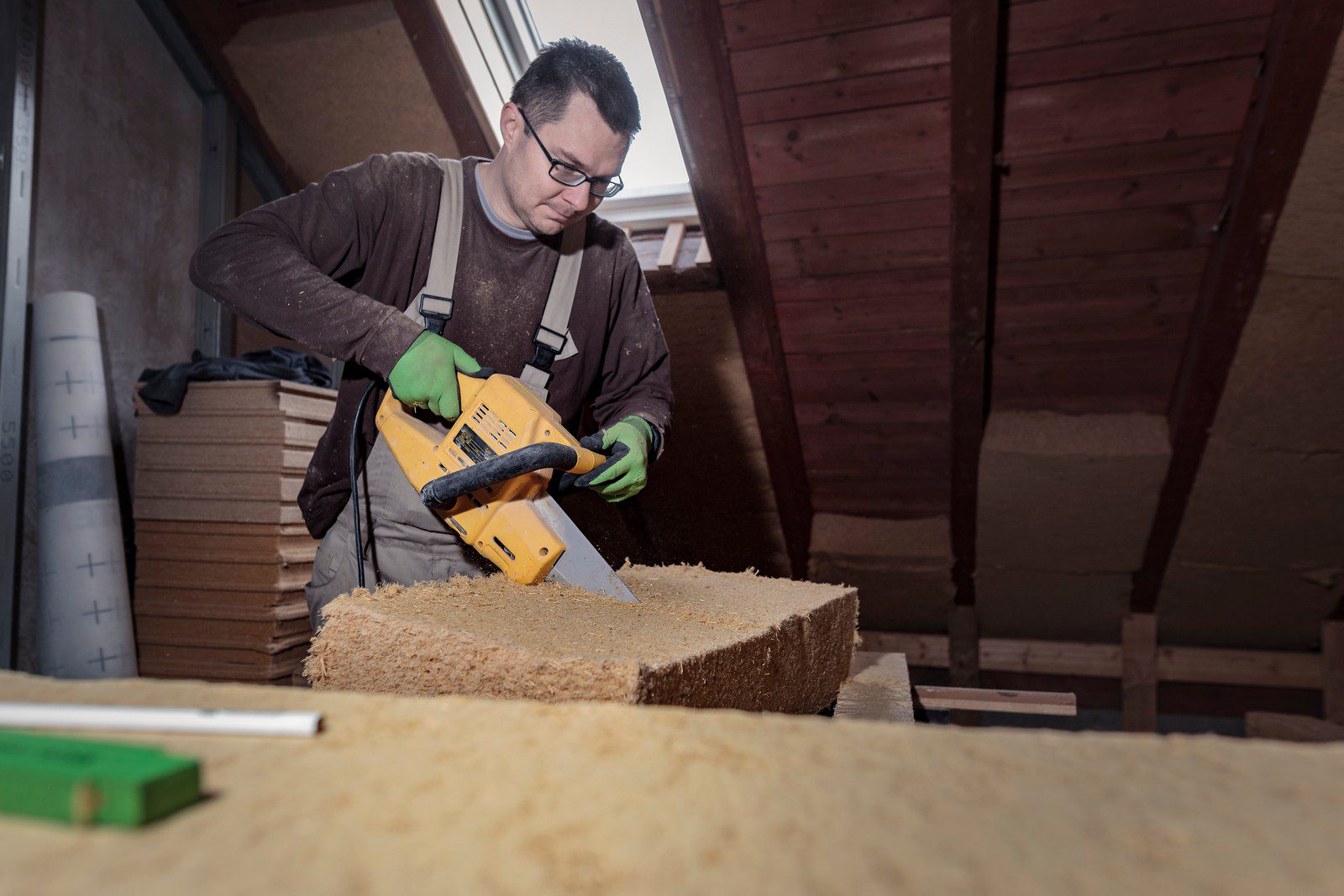 The image shows a person engaged in carpentry or construction work. The individual is using a yellow electric hand saw to cut a piece of wood. The person is wearing safety glasses, gloves for protection, and a brown long-sleeve shirt. The surrounding area looks like an attic or an unfinished room with visible insulation material, wooden beams, and planks of wood stacked nearby. There's a spirit level lying on the insulation, indicating that precision work is likely being done. Sawdust is visible, which indicates that cutting work has been ongoing.