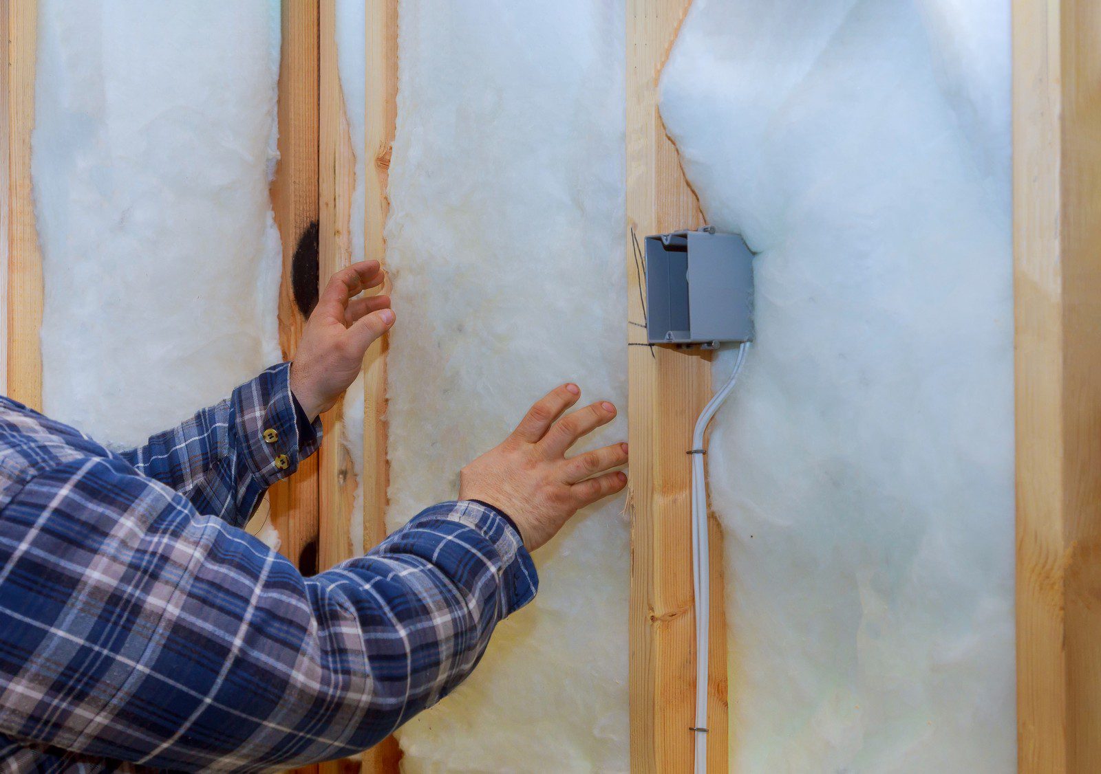 The image shows an individual installing insulation between wooden studs in a wall. The person is wearing a plaid, long-sleeved shirt and is pressing the white fibrous insulation material into place. On the right-hand side, there is an electrical junction box with conduits emerging from it, which indicates that electrical work is either planned or partially completed. This is a common step in constructing or renovating a building to ensure energy efficiency and thermal comfort.