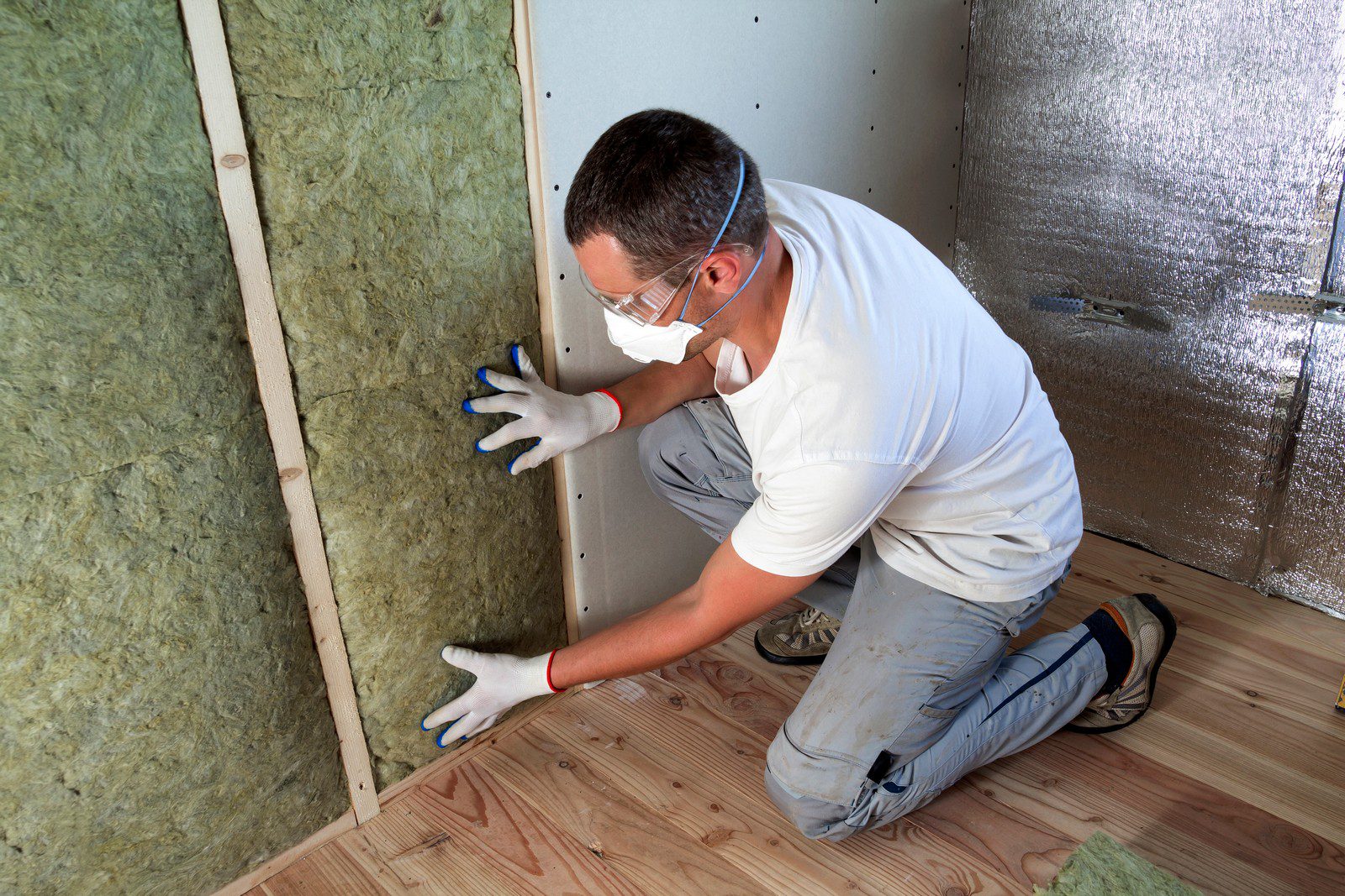 The image shows a person in the process of installing insulation in a building structure. The worker is wearing protective equipment that includes safety goggles, a dust mask, and gloves to ensure safety during the installation process. Behind the drywall and the framing studs, you can see fibrous insulation material, likely mineral wool or fiberglass, which is used to improve thermal performance and soundproofing of the walls. On the right side, there is a reflective foil barrier that may be used as an additional insulative layer or vapour barrier. The person is kneeling on a wooden subfloor, and there appears to be another piece of insulation material on the floor in the foreground. The image captures a common scene in construction and renovation settings where insulation is a critical component for energy efficiency.