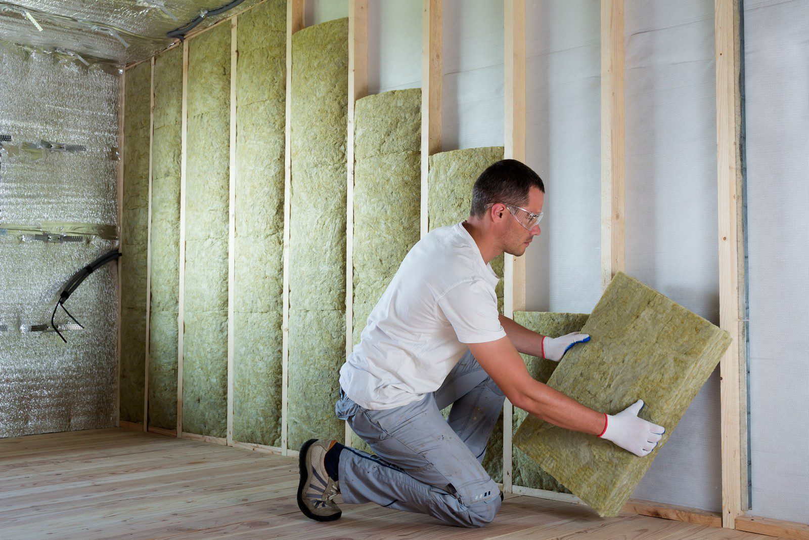 This image depicts a person engaged in the process of insulating a wall. The individual is wearing safety glasses, gloves, and casual work clothes, and is in the act of fitting a piece of mineral wool insulation between the wooden studs of a wall. The wall already has some reflective foil installed, which serves as a vapour barrier, and several pieces of insulation have been placed between other studs. The floor is wooden, and the overall setting suggests that this is a construction or renovation site where insulation is being installed for thermal efficiency and comfort.