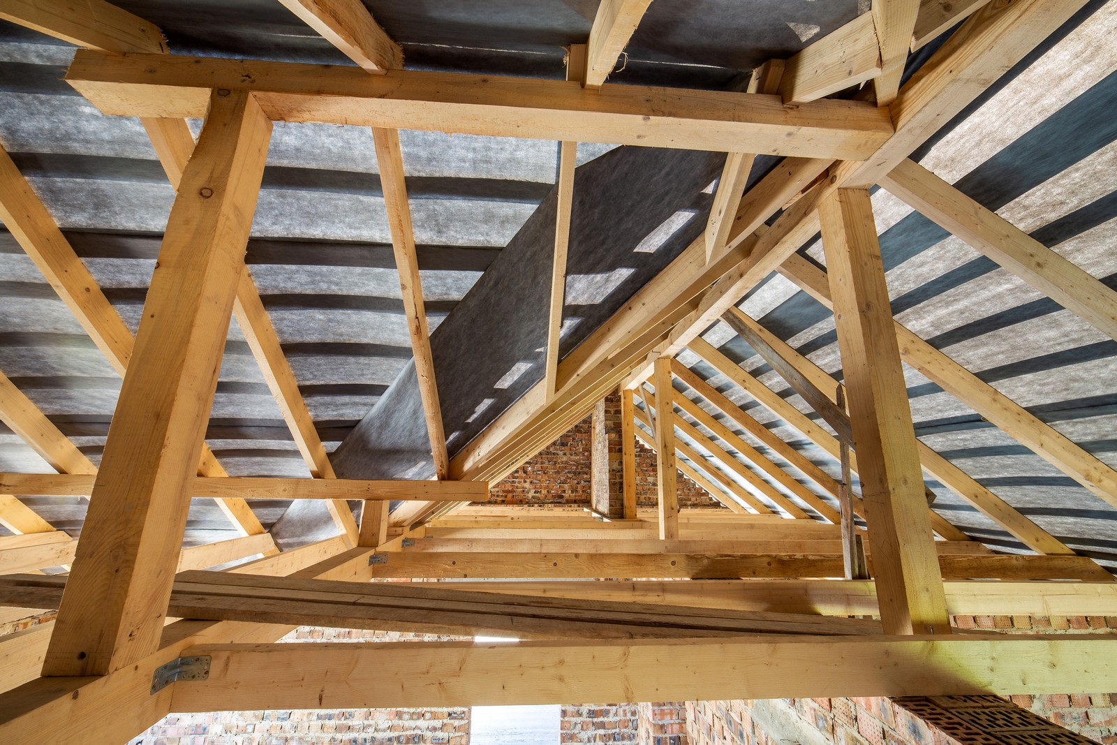 The image shows the wooden framework of a roof under construction. You can see the wooden rafters and beams, which provide the structural support for the roof. There is also some foil insulation material visible between the rafters, likely for thermal insulation purposes. In the background, a brick wall can be observed, suggesting this is part of a building that may either be undergoing renovations or in the process of being built. Overall, the image captures the intricacies of wooden roof structure and building construction.