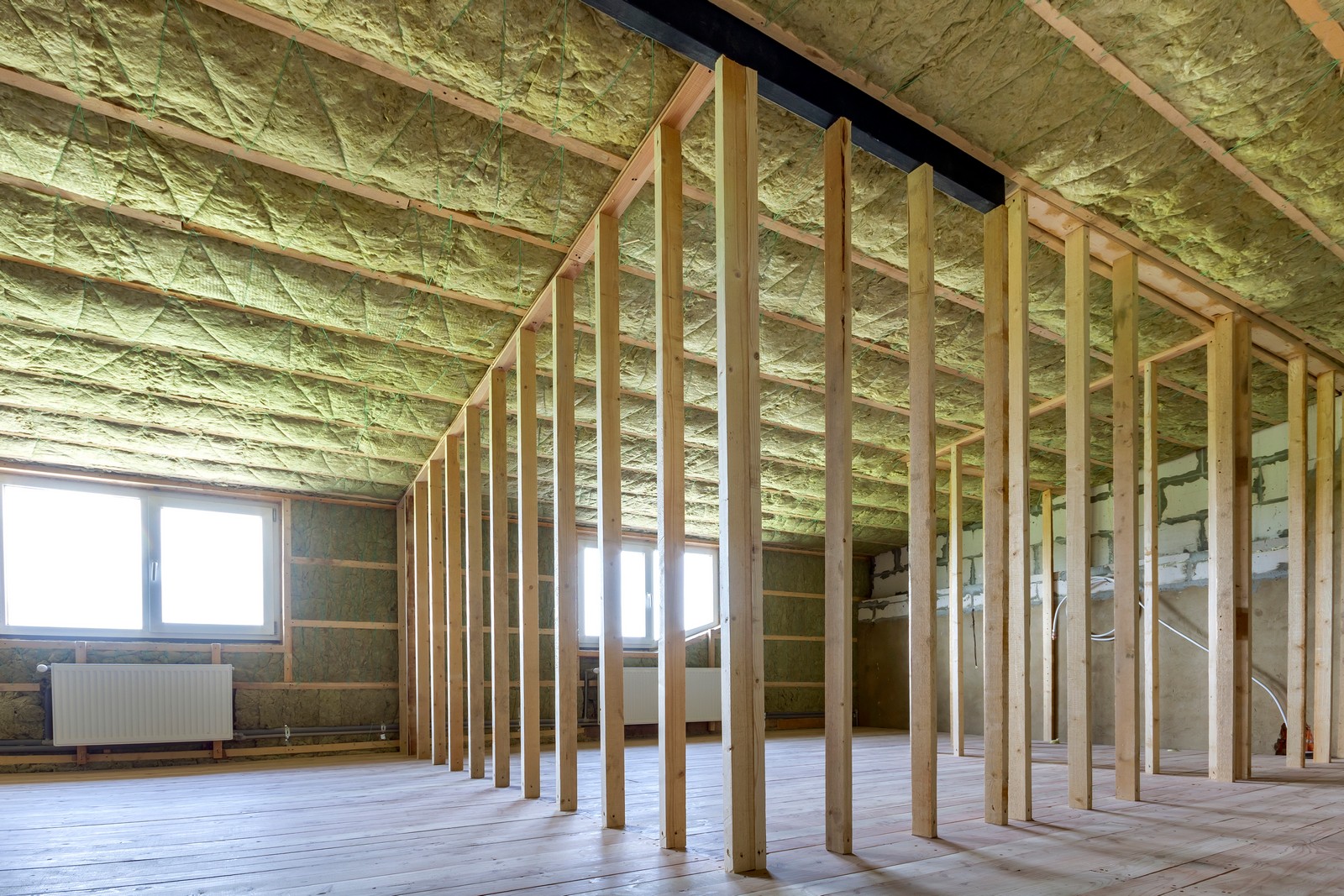 The image shows an attic under construction or renovation. The ceiling has been insulated with fibrous insulation material, often used for thermal insulation to keep heat from escaping during the winter and to keep the space cool during the summer. You can also see a wooden stud framework, which likely outlines where walls or additional rooms will be created within the attic space. The construction uses wooden planks for the floor and vertical structural timber pieces that are part of the stud framework. There is a radiator near the window, suggesting that the space is intended to be habitable. The windows indicate that natural light is available in the space. Additionally, the presence of vapour barriers on the insulation shows attention to moisture control, which is crucial in preventing mould and structural damage in an insulated space.