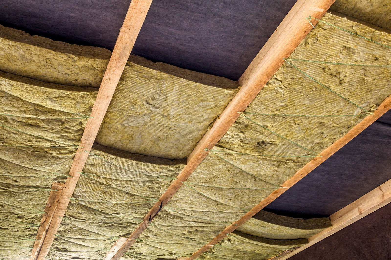 The image shows a close-up view of building insulation materials. There are layers of fiberglass insulation wedged between wooden beams, typically referred to as joists. The insulation's yellow colour and fibrous texture are clearly visible, and it appears to be unfaced, meaning there's no paper or foil facing attached to it. The dark area could be a vapour barrier or simply a shadowed area beyond the insulation. This kind of material is commonly used to insulate buildings to retain heat or cool air, enhancing energy efficiency and comfort.