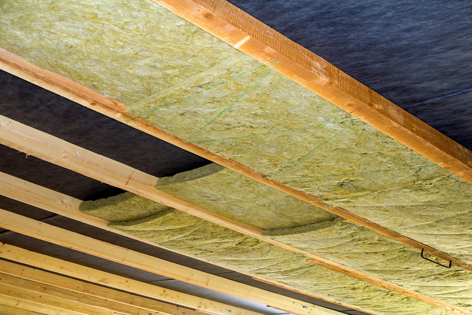 The image shows the underside of a roof structure with insulation material installed between the joists. The insulation appears to be fiberglass, recognizable by its fibrous texture and yellow colour. The wooden beams, or joists, provide structural support for the roof and also act as a framework for the insulation material. There is also a black layer visible above the insulation, which could be a vapour barrier, designed to prevent moisture from passing through. It looks like a typical construction or renovation site where the roof of a building is being insulated for energy efficiency. One of the bays between the joists is missing insulation, revealing the dark barrier beneath. There's also a wire or cable hanging down, which might be part of the electrical installation.